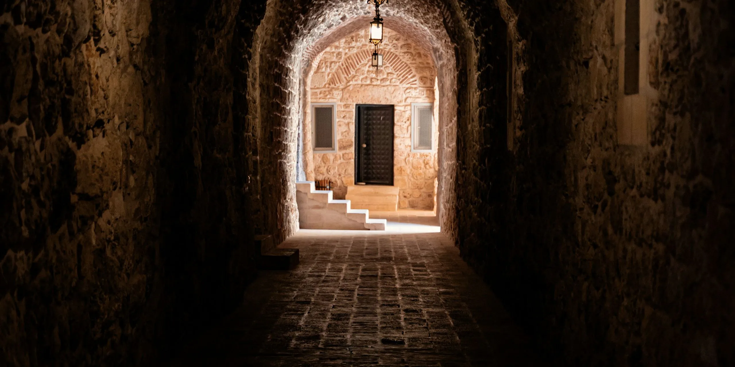 Vaulted stone passage lit by a lantern, opening onto a door and a sunlit staircase